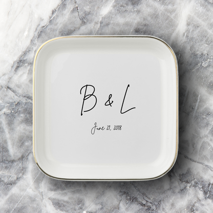 A square white ceramic dish with rounded corners, featuring a thin gold rim and personalized black script text reading 'B & L' and the date 'June 21, 2018', set on a gray and white marble surface.