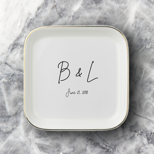 A square white ceramic dish with rounded corners, featuring a thin gold rim and personalized black script text reading 'B & L' and the date 'June 21, 2018', set on a gray and white marble surface.