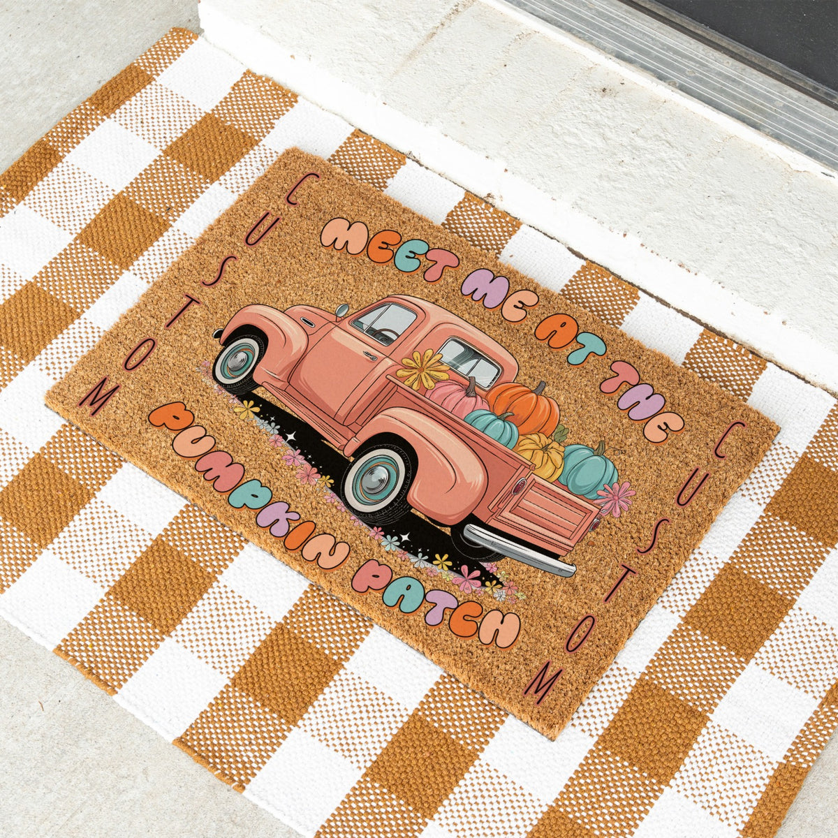 A rectangular doormat with a rustic autumn design, featuring a pink pickup truck loaded with pumpkins and the phrase 'MEET ME AT THE PUMPKIN PATCH' on a tan background. The doormat is styled on a brown and white checkered blanket.