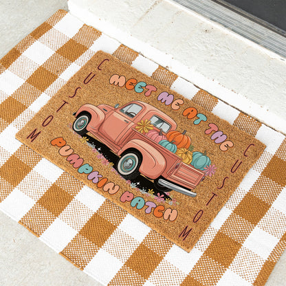 A rectangular doormat with a rustic autumn design, featuring a pink pickup truck loaded with pumpkins and the phrase 'MEET ME AT THE PUMPKIN PATCH' on a tan background. The doormat is styled on a brown and white checkered blanket.