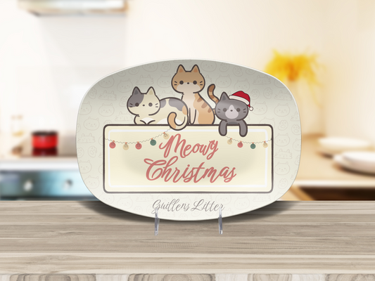 A decorative ceramic platter with rounded corners, featuring three cartoon cats above the words 'Meowy Christmas' in pink script, displayed on a small stand on a wooden surface with a blurred kitchen background.