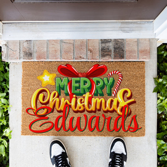 A personalized coir doormat with a festive Christmas design, shown on a concrete porch with a person's shoes visible at the bottom. The doormat features the text 'MERRY Christmas & Edwards' in red, yellow, and green, decorated with a red bow, candy cane, and star.