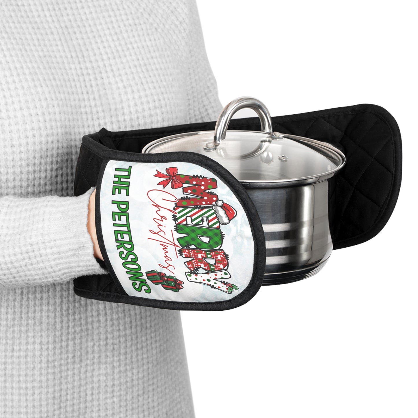 A person wearing a light gray waffle-knit sweater holds a stainless steel pot with a glass lid using a black quilted oven mitt. The mitt features a white front panel with a Christmas-themed design that is custom and reads 'The Petersons Merry Christmas' in green and red lettering with red gift boxes.