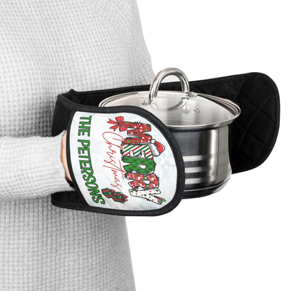 A person wearing a light gray waffle-knit sweater holds a stainless steel pot with a glass lid using a black quilted oven mitt. The mitt features a white front panel with a Christmas-themed design that is custom and reads 'The Petersons Merry Christmas' in green and red lettering with red gift boxes.