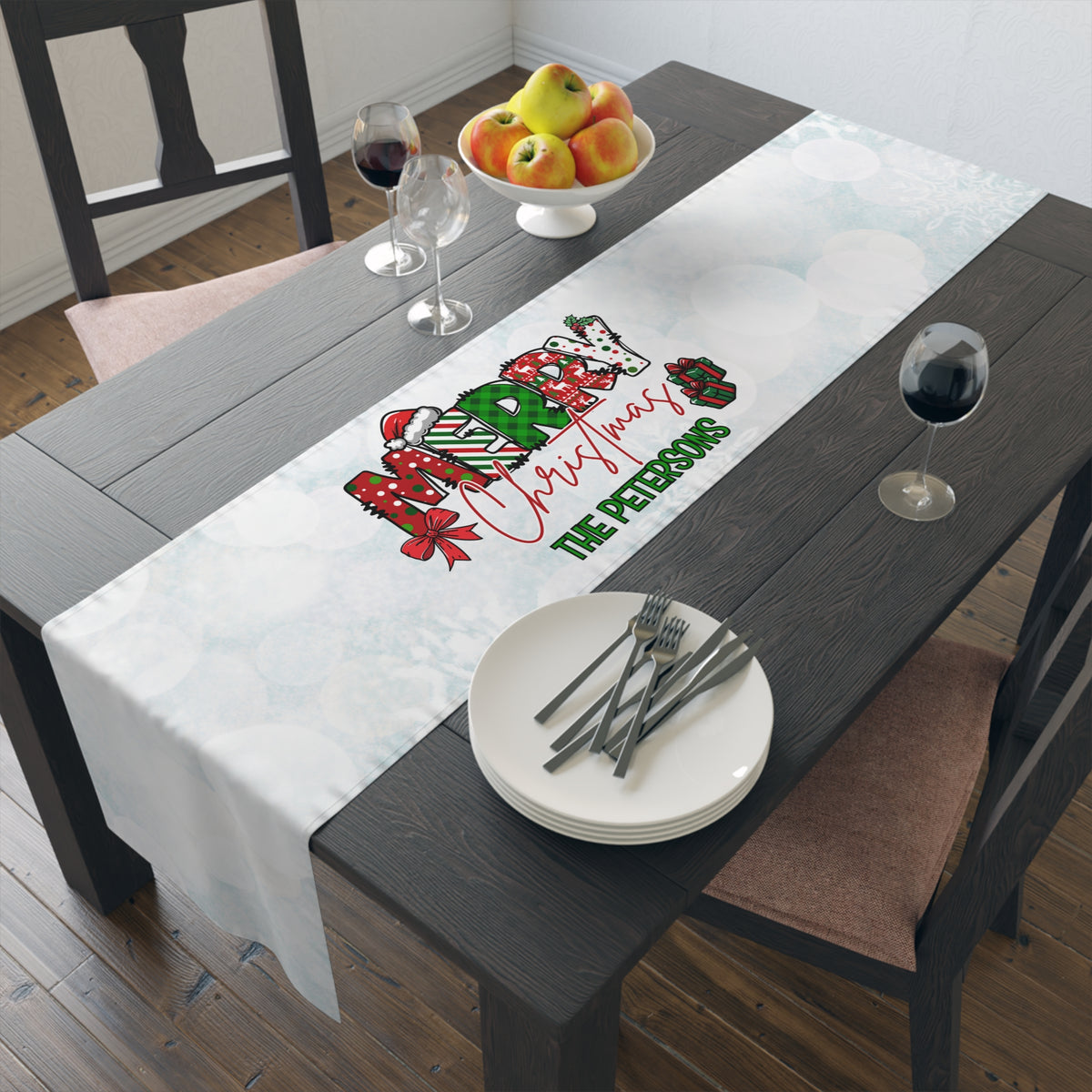 A personalized white Christmas table runner with a festive graphic and the text 'The Petersons' is displayed on a dark wooden dining table. The table is styled with white plates, silver cutlery, wine glasses, and a bowl of apples.