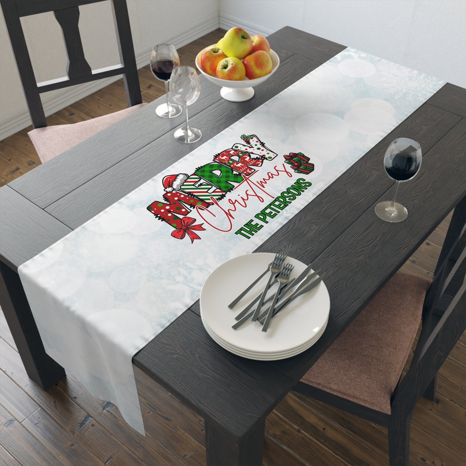 A personalized white Christmas table runner with a festive graphic and the text 'The Petersons' is displayed on a dark wooden dining table. The table is styled with white plates, silver cutlery, wine glasses, and a bowl of apples.
