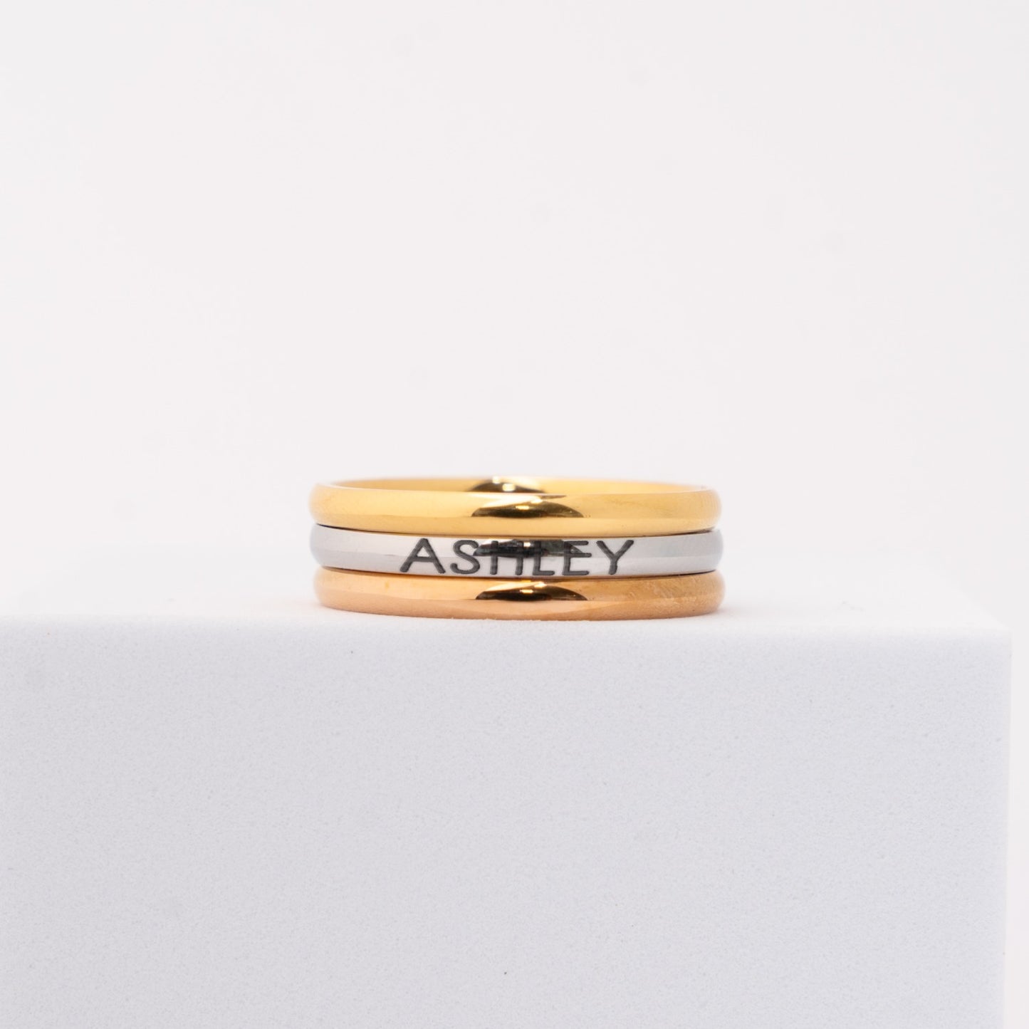 3 personalized rings featuring gold, rose gold and silver bands stacked together, with the name 'ASHLEY' engraved on the silver band, displayed on a white block against a white background.