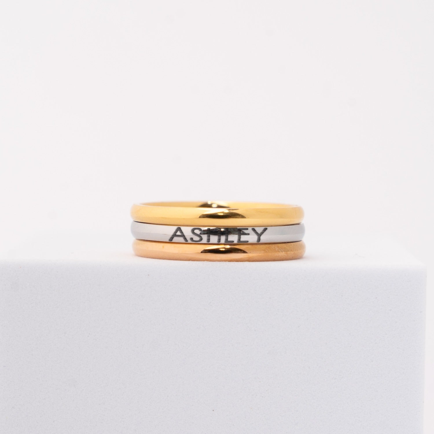 3 personalized rings featuring gold, rose gold and silver bands stacked together, with the name 'ASHLEY' engraved on the silver band, displayed on a white block against a white background.
