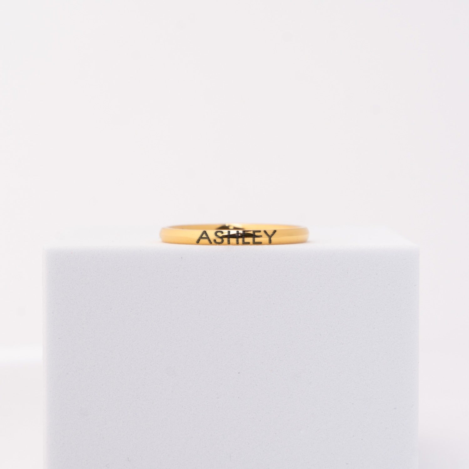 A thin yellow gold band ring engraved with the name 'ASHLEY' in black lettering, displayed on a white cube against a white background.