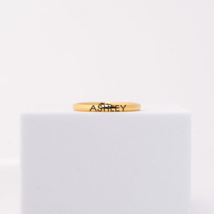 A thin yellow gold band ring engraved with the name 'ASHLEY' in black lettering, displayed on a white cube against a white background.