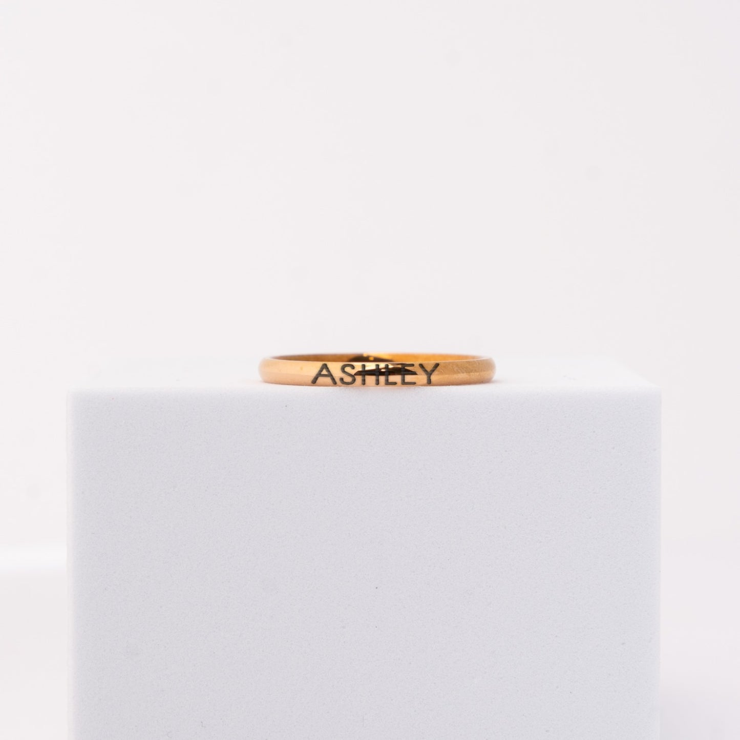 A thin rose gold band ring engraved with the name 'ASHLEY' in black lettering, displayed on a white cube against a white background.