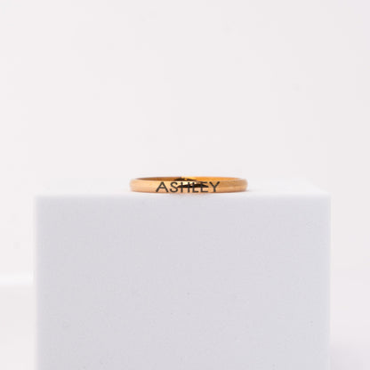 A thin rose gold band ring engraved with the name 'ASHLEY' in black lettering, displayed on a white cube against a white background.