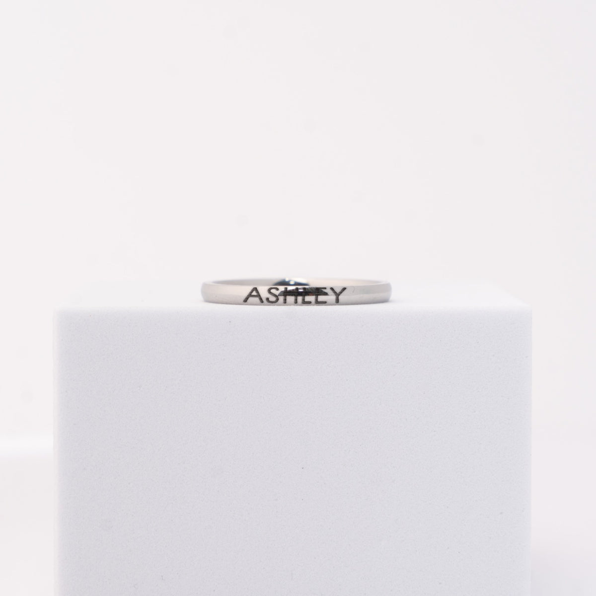 A silver band ring engraved with the name 'ASHLEY'  displayed on a white cube against a white background.