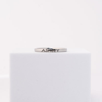 A silver band ring engraved with the name 'ASHLEY'  displayed on a white cube against a white background.