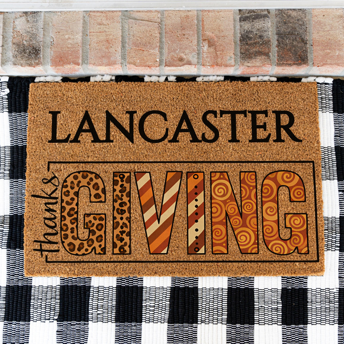 A rectangular coir doormat with the words “LANCASTER thanksGIVING”  placed in front of a brick threshold and a black-and-white checkered rug. The letters in “GIVING” feature decorative patterns including leopard print, stripes, and swirls in warm brown, orange, tan, and black tones.