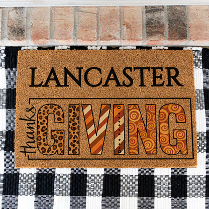 A rectangular coir doormat with the words “LANCASTER thanksGIVING”  placed in front of a brick threshold and a black-and-white checkered rug. The letters in “GIVING” feature decorative patterns including leopard print, stripes, and swirls in warm brown, orange, tan, and black tones.