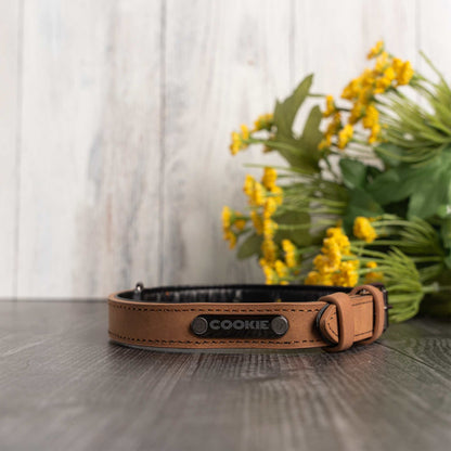 A brown leather dog collar with a black interior and the name 'COOKIE' printed on the front, resting on a dark wooden surface with blurred yellow flowers in the background.