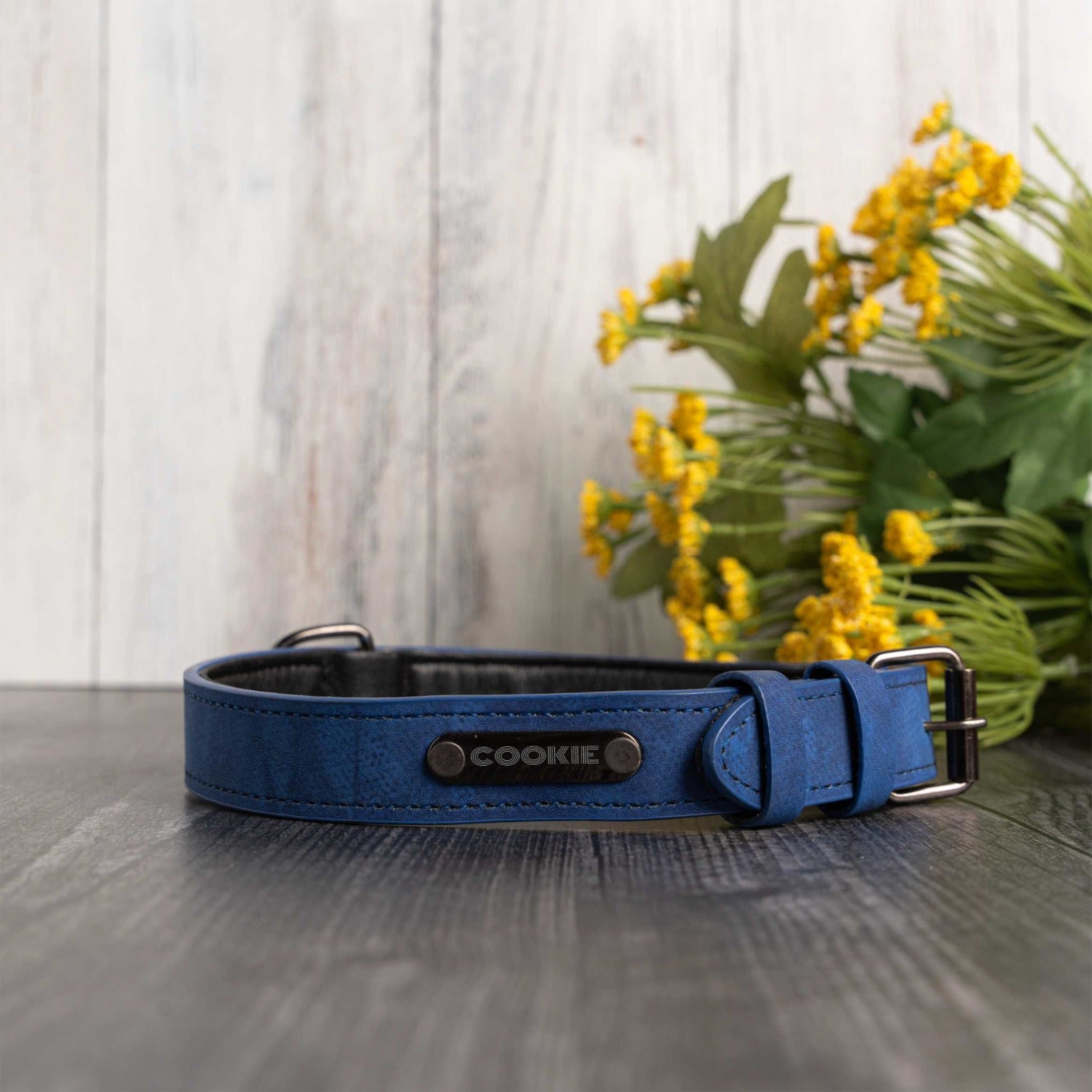 A blue leather dog collar with a silver buckle and a central metal plate engraved with the name 'COOKIE', displayed on a dark wood surface with yellow flowers in the background.