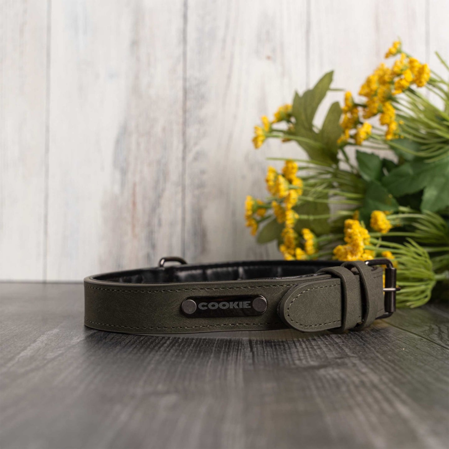 An olive green leather dog collar with a black interior and a silver-tone buckle, with custom name cookie on front resting on a dark wooden surface with yellow flowers in the background.