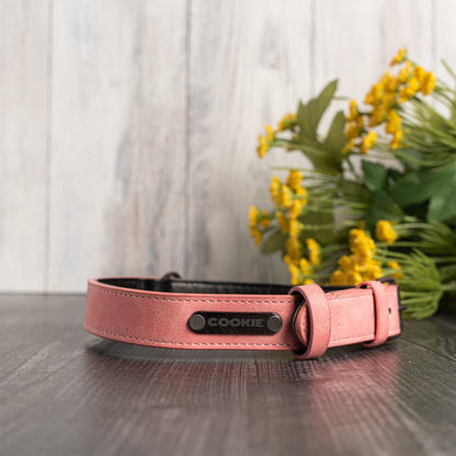 A pink leather dog collar with a black metal plate engraved with the name 'COOKIE', resting on a dark wooden surface with yellow flowers in the background.