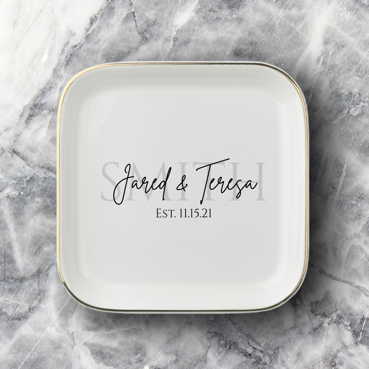 A square white ceramic dish with rounded corners, featuring a thin gold rim and personalized black script text reading 'Jared & Teresa' with personalized last name behind that reading 'SMITH' and the date 'Est. 11.15.21' below that, set on a gray and white marble surface.