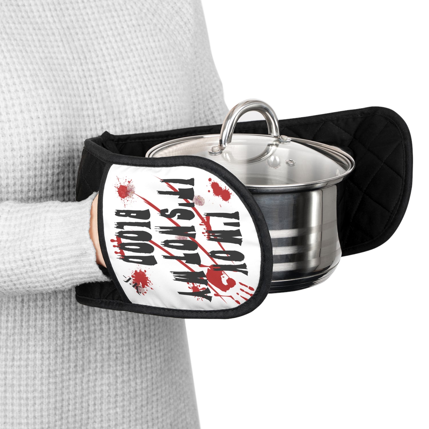 A person wearing a gray waffle-knit sweater holds a black quilted oven mitt with a white graphic that reads 'I'M OK IT'S NOT MY BLOOD' in black text with red blood splatter details. and a custom text initials on the bottom. A stainless steel pot with a glass lid sits inside the mitt.