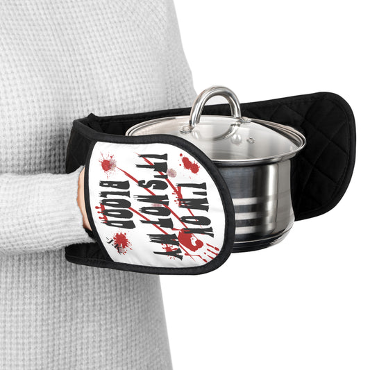A person wearing a gray waffle-knit sweater holds a black quilted oven mitt with a white graphic that reads 'I'M OK IT'S NOT MY BLOOD' in black text with red blood splatter details. and a custom text initials on the bottom. A stainless steel pot with a glass lid sits inside the mitt.