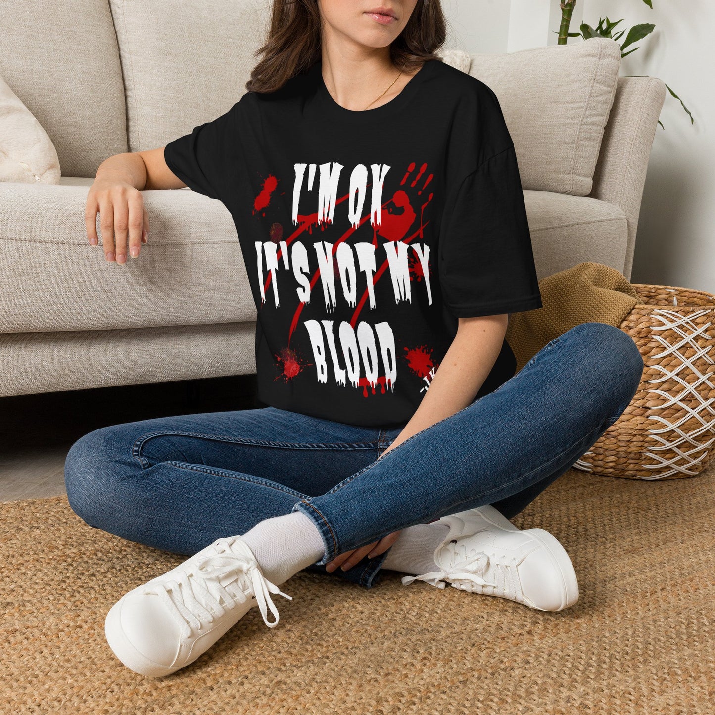 A woman sitting cross-legged on a woven rug in front of a beige sofa, wearing a black graphic T-shirt with distressed white text reading 'I'M OK IT'S NOT MY BLOOD' over a red splatter design, paired with blue jeans and white sneakers.