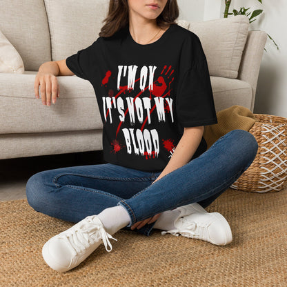 A woman sitting cross-legged on a woven rug in front of a beige sofa, wearing a black graphic T-shirt with distressed white text reading 'I'M OK IT'S NOT MY BLOOD' over a red splatter design, paired with blue jeans and white sneakers.