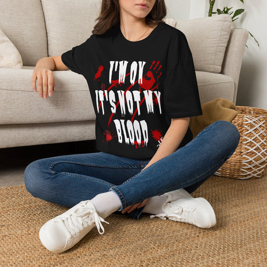 A woman sitting cross-legged on a woven rug in front of a beige sofa, wearing a black graphic T-shirt with distressed white text reading 'I'M OK IT'S NOT MY BLOOD' over a red splatter design, paired with blue jeans and white sneakers.