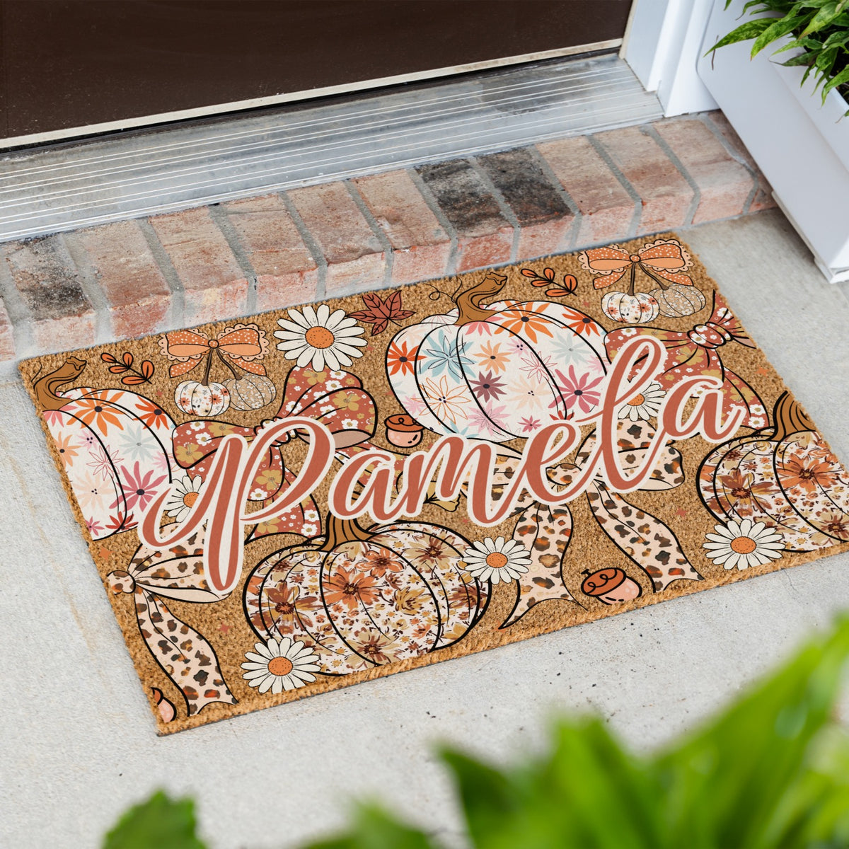 A personalized coir doormat with an autumn pumpkin and daisy pattern, featuring the name 'Pamela' in script, placed on a concrete porch step in front of a brick threshold.