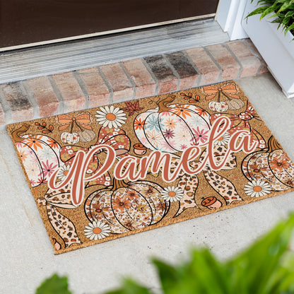 A personalized coir doormat with an autumn pumpkin and daisy pattern, featuring the name 'Pamela' in script, placed on a concrete porch step in front of a brick threshold.