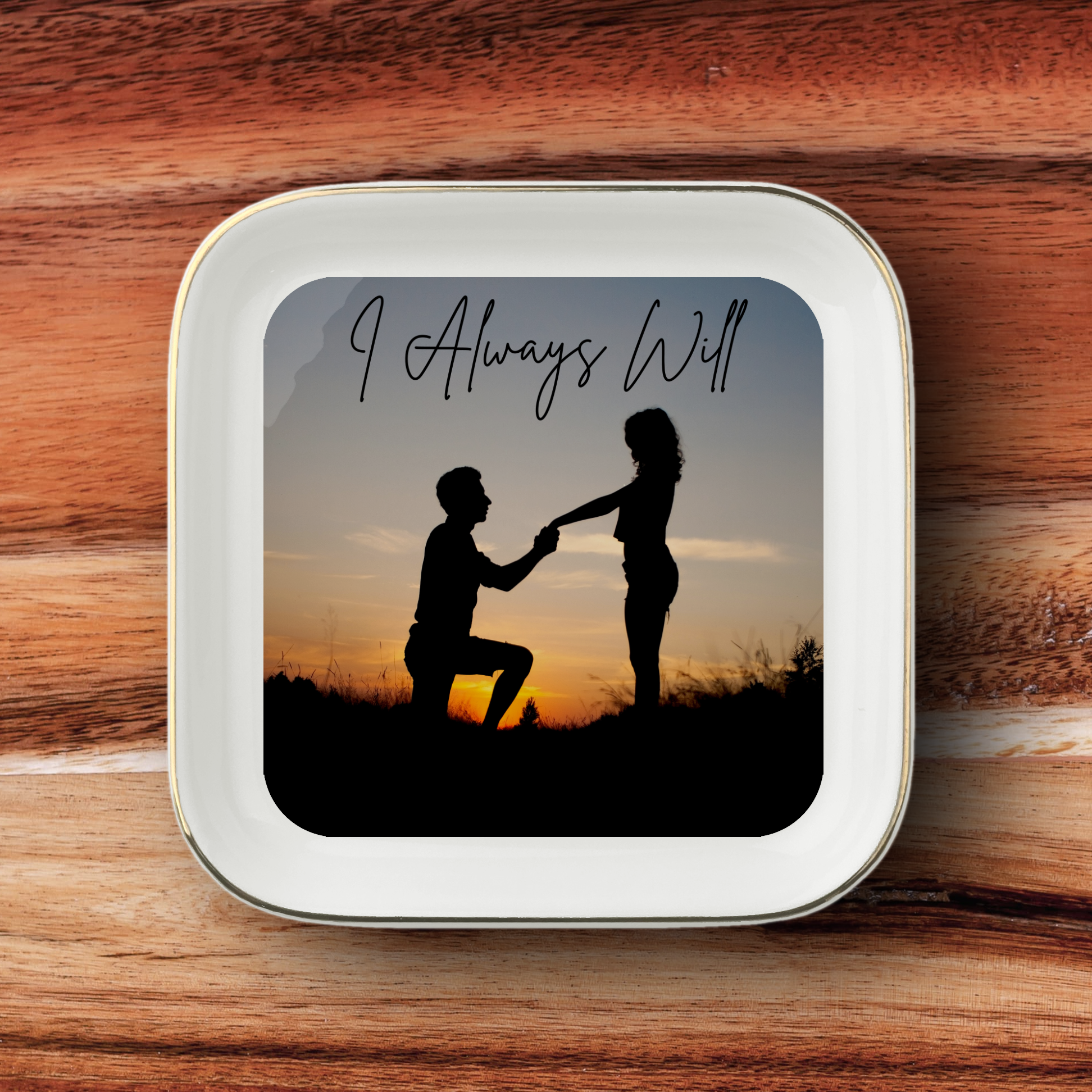 A square white ceramic jewelry dish with rounded corners and a gold metallic rim, featuring a printed square graphic of a silhouette couple holding hands at sunset with the text 'I Always Will'. The jewelry dish is placed on a rustic wooden surface.