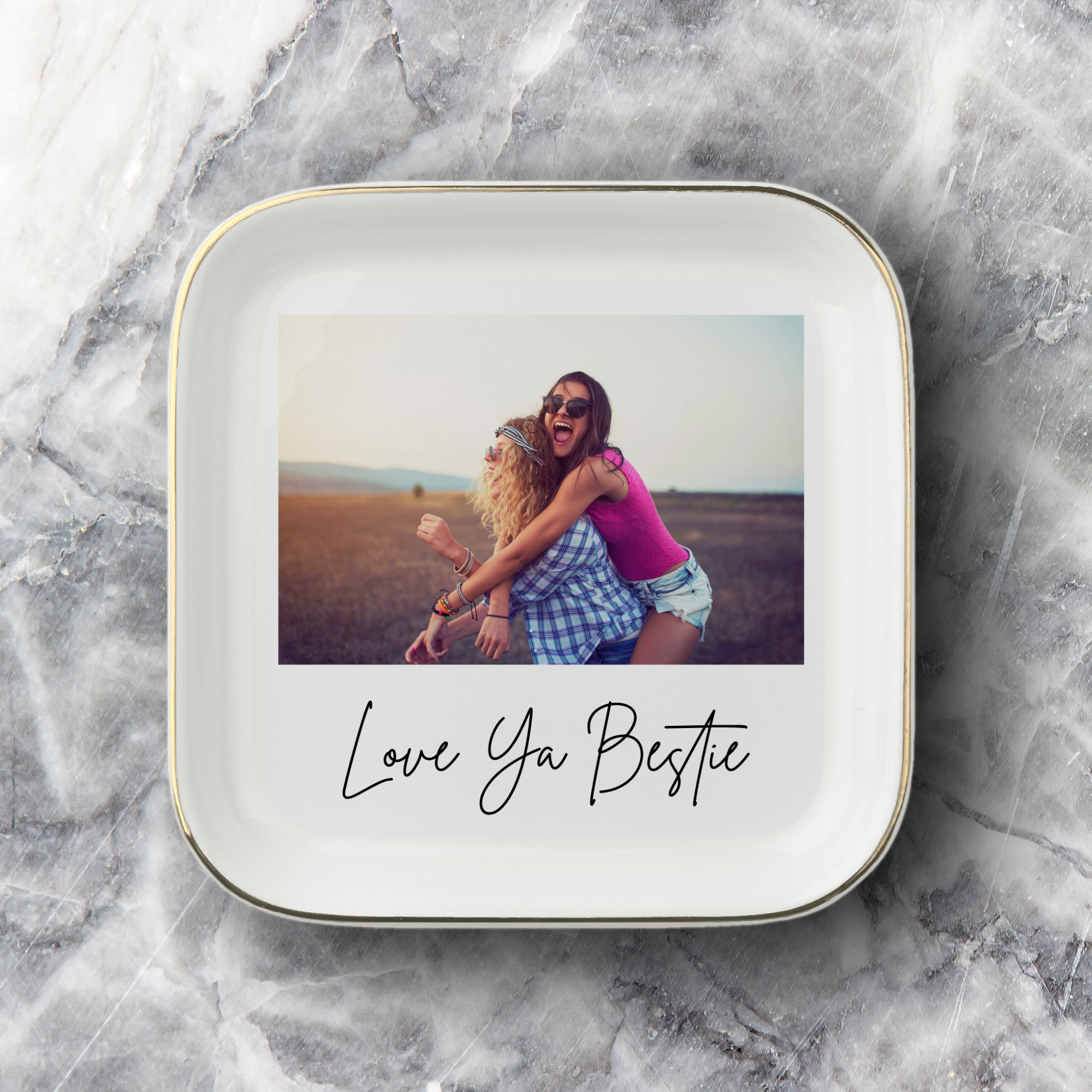 A square white ceramic photo jewelry dish with rounded corners, featuring a printed photo of two women hugging and the text 'Love Ya Bestie' in black script underneath photo. The jewelry dish is set on a gray and white marble surface.
