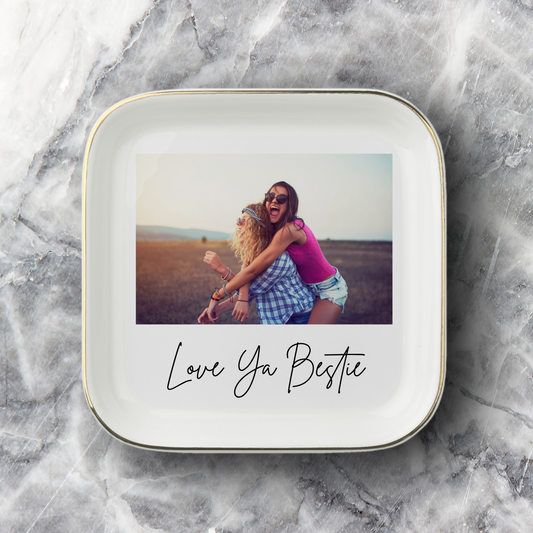 A square white ceramic photo jewelry dish with rounded corners, featuring a printed photo of two women hugging and the text 'Love Ya Bestie' in black script underneath photo. The jewelry dish is set on a gray and white marble surface.