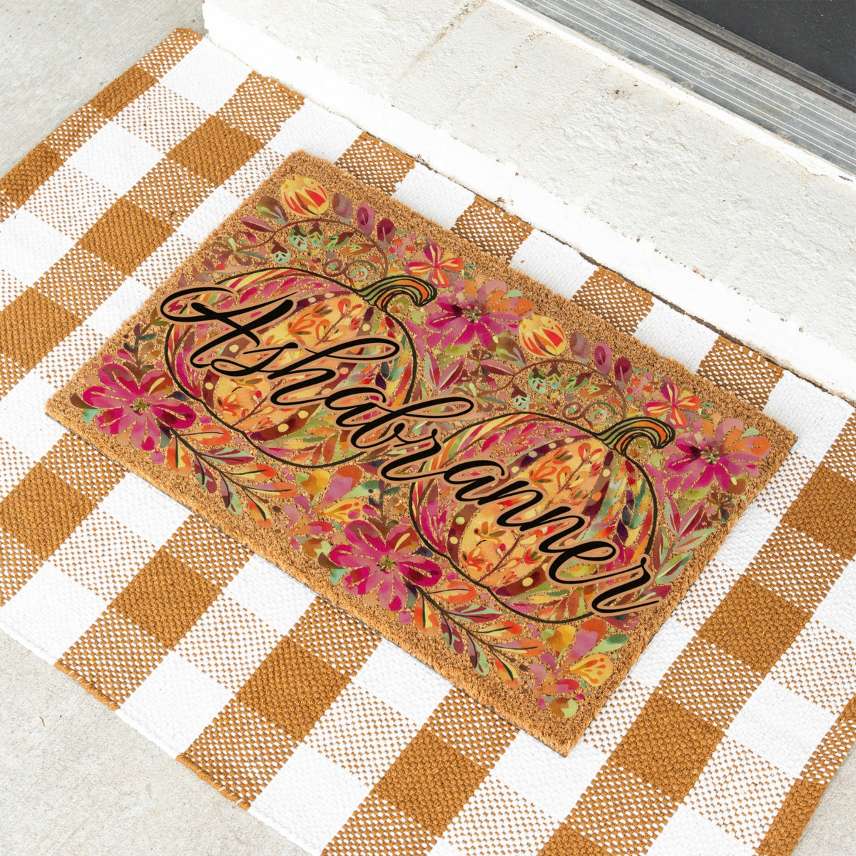 A rectangular doormat with a fall harvest design, featuring two pumpkins and the name 'Ashbranner' in black script. The doormat is placed on a white and brown gingham checkered mat on a light concrete floor.