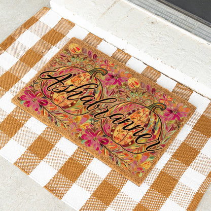 A rectangular doormat with a fall harvest design, featuring two pumpkins and the name 'Ashbranner' in black script. The doormat is placed on a white and brown gingham checkered mat on a light concrete floor.