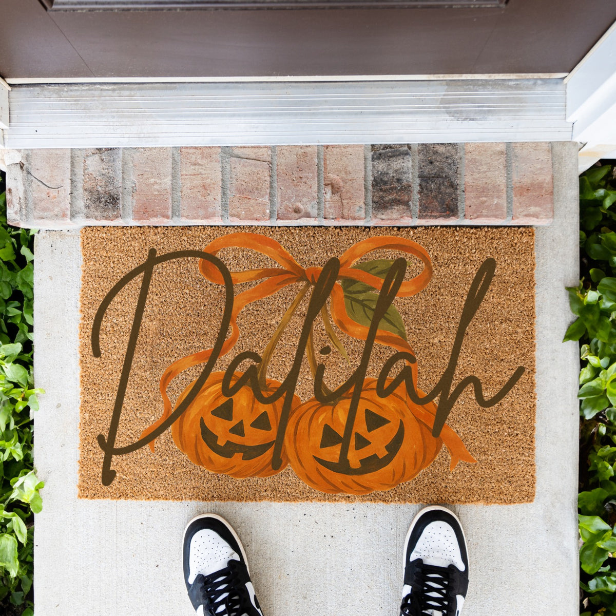 A rectangular coir doormat with a personalized name, 'Dalilah', written in dark brown script over an illustration of two orange pumpkins hanging like cherries, shown on a front porch with a person's shoes visible at the bottom.