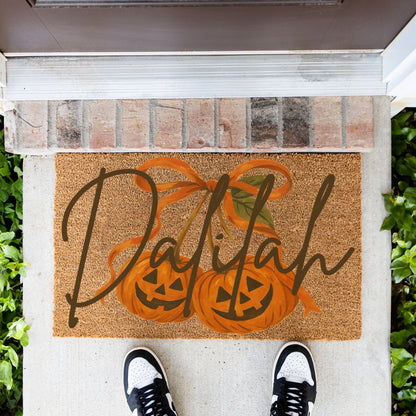 A rectangular coir doormat with a personalized name, 'Dalilah', written in dark brown script over an illustration of two orange pumpkins hanging like cherries, shown on a front porch with a person's shoes visible at the bottom.