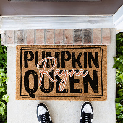 A coir doormat with the text 'PUMPKIN Rayna QUEEN' in black and orange script, placed at a front door threshold. A person's feet in black and white sneakers are visible at the bottom of the frame, with brick steps, greenery, and a white threshold visible.