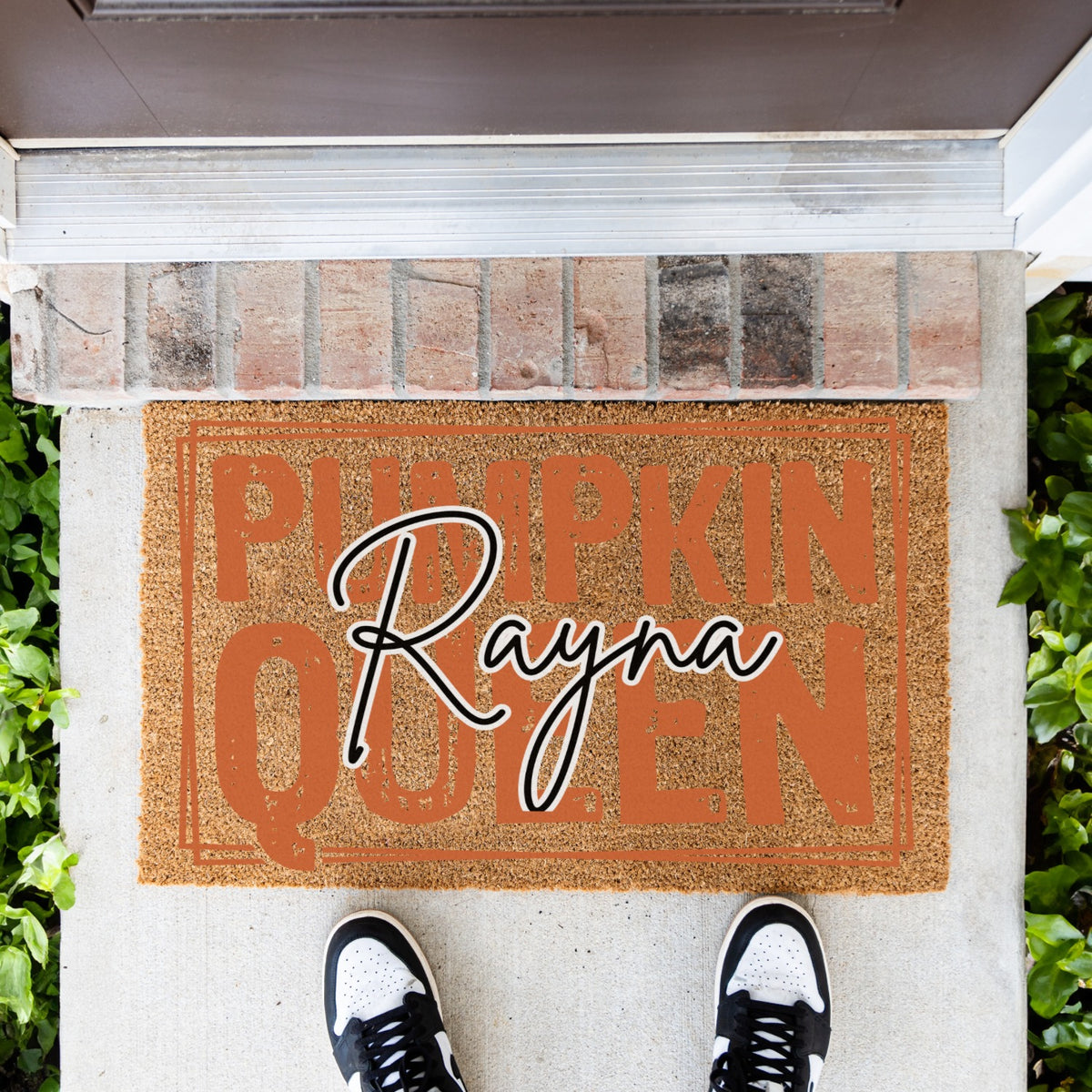 A doormat with orange design that says pumpkin queen with the custom name rayna in black and white. the doormat is on the concete infront of the door and some shoes are infront of it