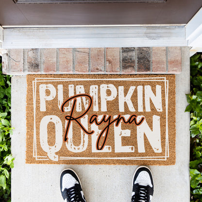 A rectangular coir doormat with the words “PUMPKIN QUEEN” in distressed white letters and a black and orange custom script overlay reading “Rayna,” placed at a front door threshold. A person’s feet in black-and-white sneakers are visible at the bottom, with brick steps, concrete, and greenery around it.