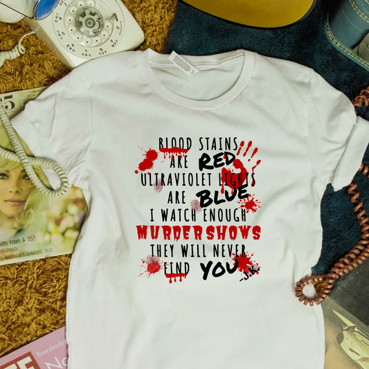 A white short-sleeve graphic T-shirt laid flat on a textured brown rug with vintage props around it. The shirt features a horror-themed text print in black and red: 'Blood stains are red, ultraviolet lights are blue, I watch enough murders shows they will never find you,' with red handprint and blood splatter graphics.