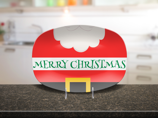 A glossy decorative platter, featuring a red upper section with a white beard, the text 'MERRY CHRISTMAS' in green on a white band, and a black lower section with a yellow belt buckle, displayed on a kitchen countertop.