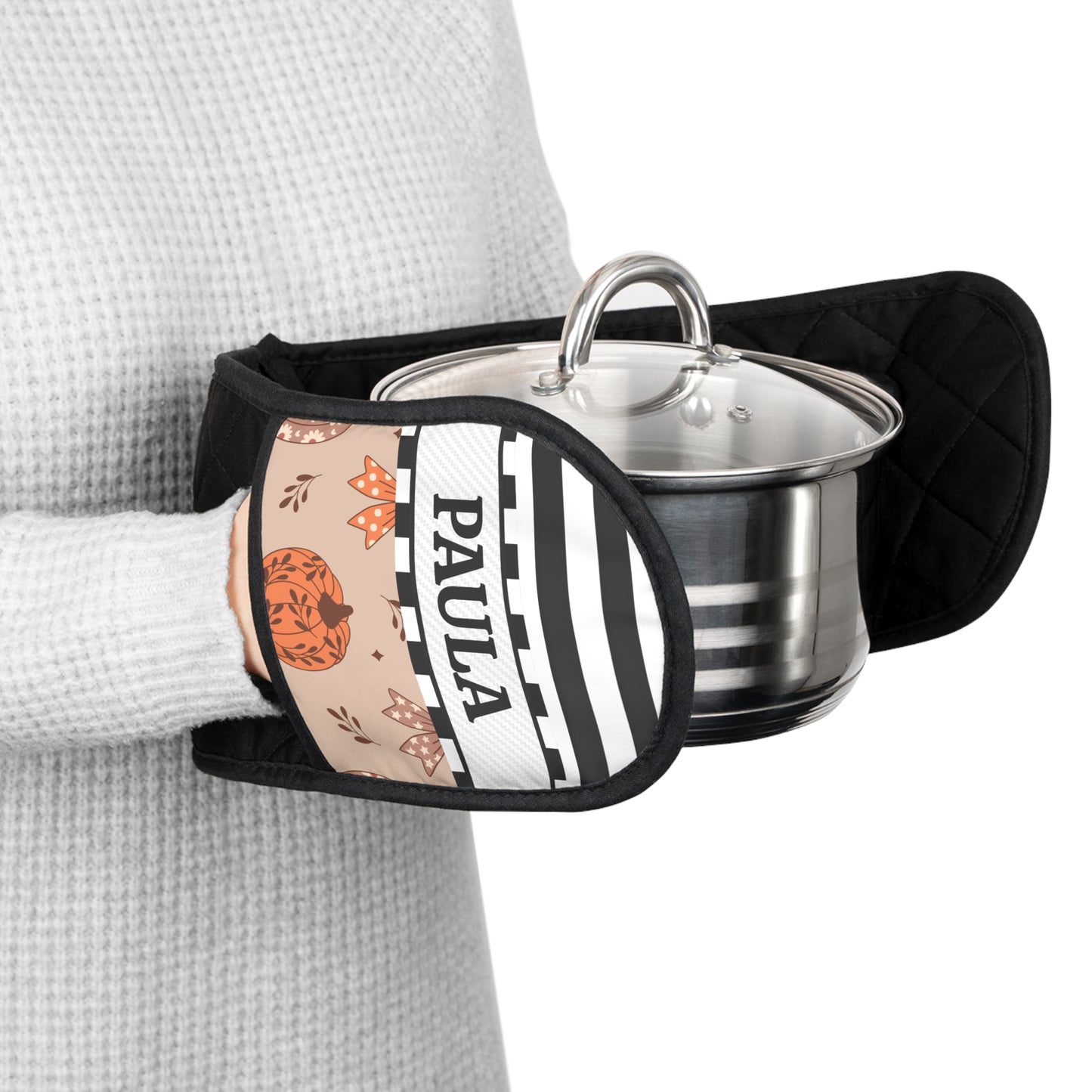 A close-up of a person wearing a light gray waffle-knit sweater and holding a black quilted pot holder with a custom printed front panel. The printed panel features black and white stripes, the name 'PAULA' in black text, and an orange pumpkin and bows illustration.