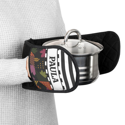 A person wearing a gray waffle-knit sweater holds a stainless steel pot with a glass lid using a black quilted oven mitt. The mitt features a custom design with the custom name 'PAULA' in black text on a white background, alongside a multicolor autumn leaf pattern.