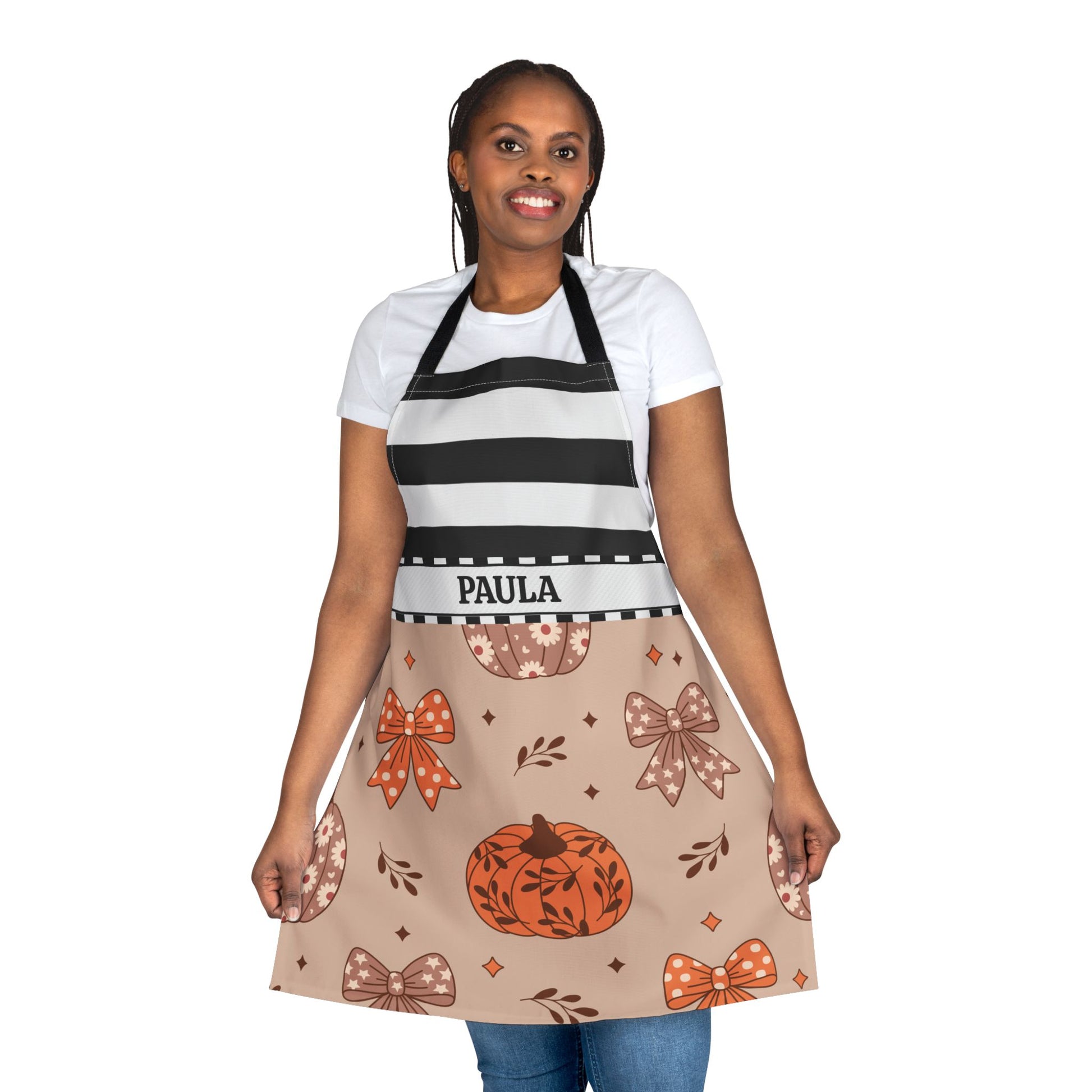 A woman modeling a personalized apron with a black and white striped top section and a beige lower section decorated with a pumpkin, bows, and leaves pattern. The name 'PAULA' is printed on the waistband.