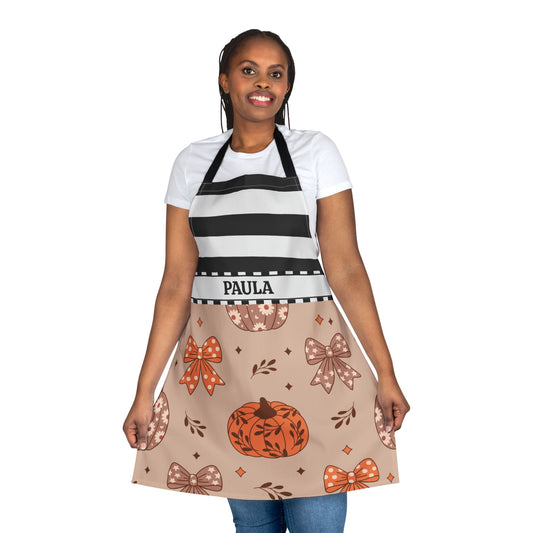 A woman modeling a personalized apron with a black and white striped top section and a beige lower section decorated with a pumpkin, bows, and leaves pattern. The name 'PAULA' is printed on the waistband.