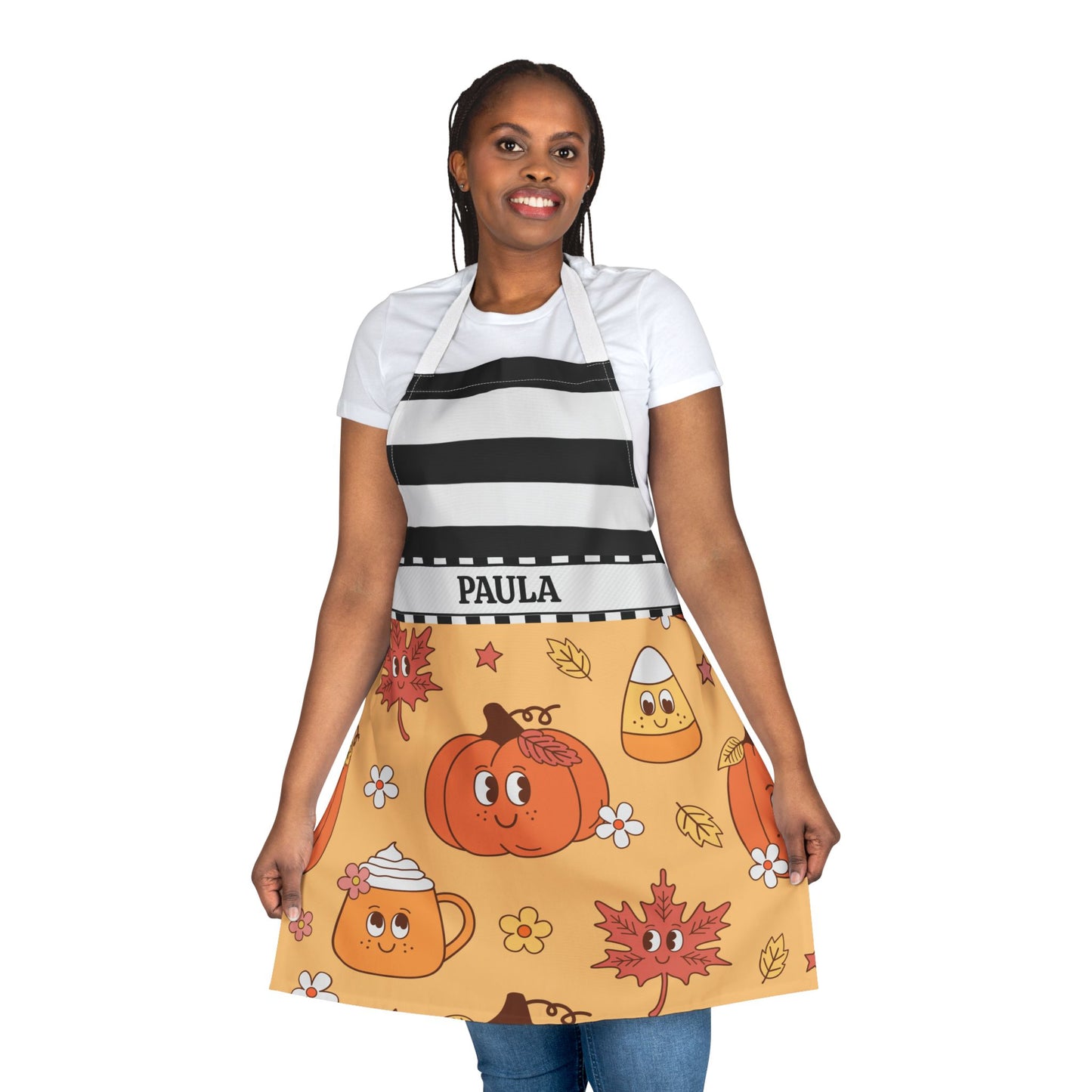 A woman modeling an apron with a black and white striped upper section and a yellowish/orangeish lower skirt featuring a cartoon pumpkin, candy corn, autumn leaves, and the name 'PAULA' on a white waistband.