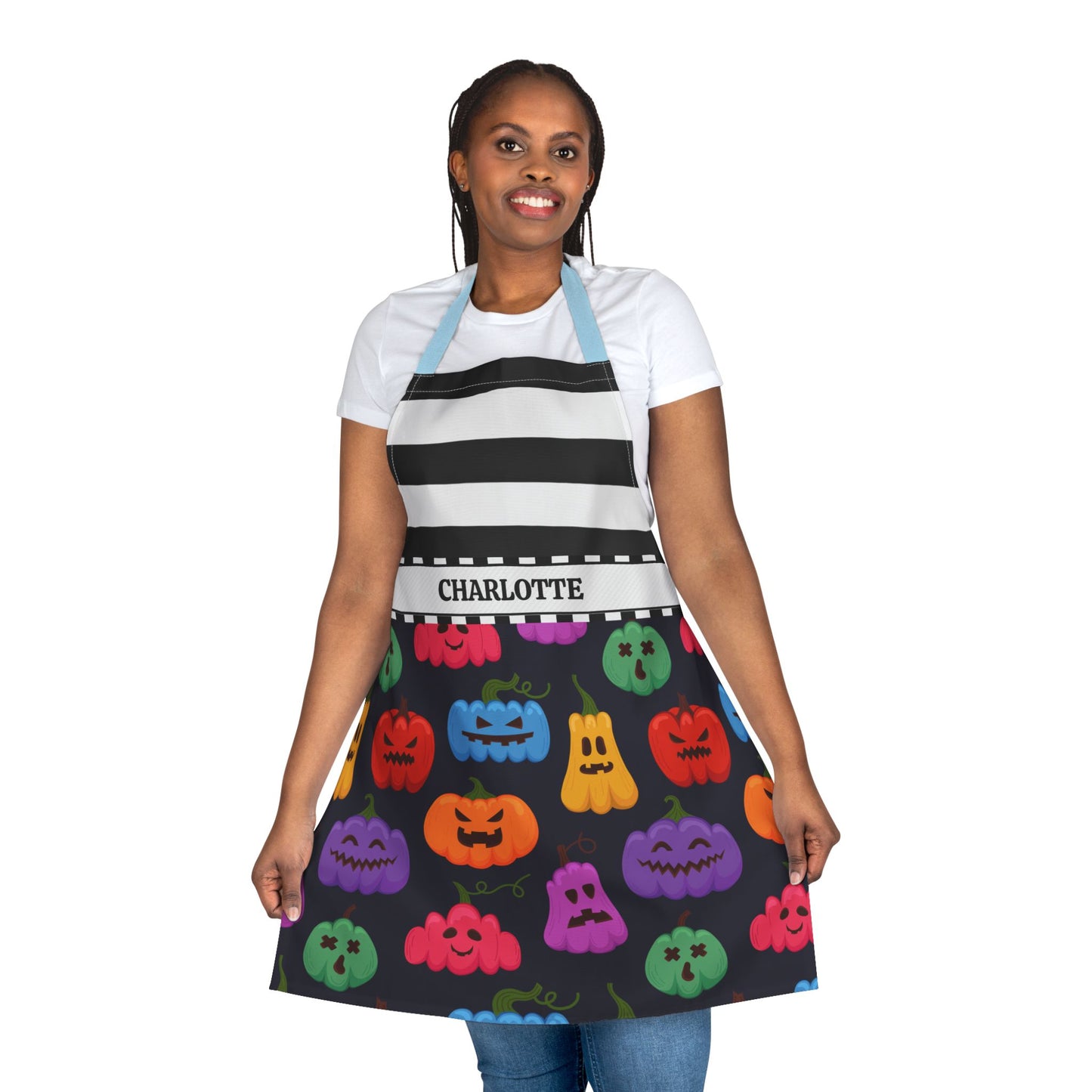 A woman modeling a black kitchen apron with a black and white striped yoke and a colorful Halloween pumpkin print skirt. The apron has a white waistband with the name 'CHARLOTTE' printed on it, and light blue neck and shoulder straps.